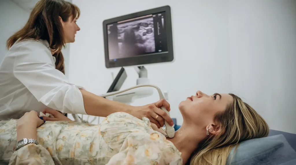 A woman lies on an examination table as an ultrasound is performed on her throat, with medical equipment in view.