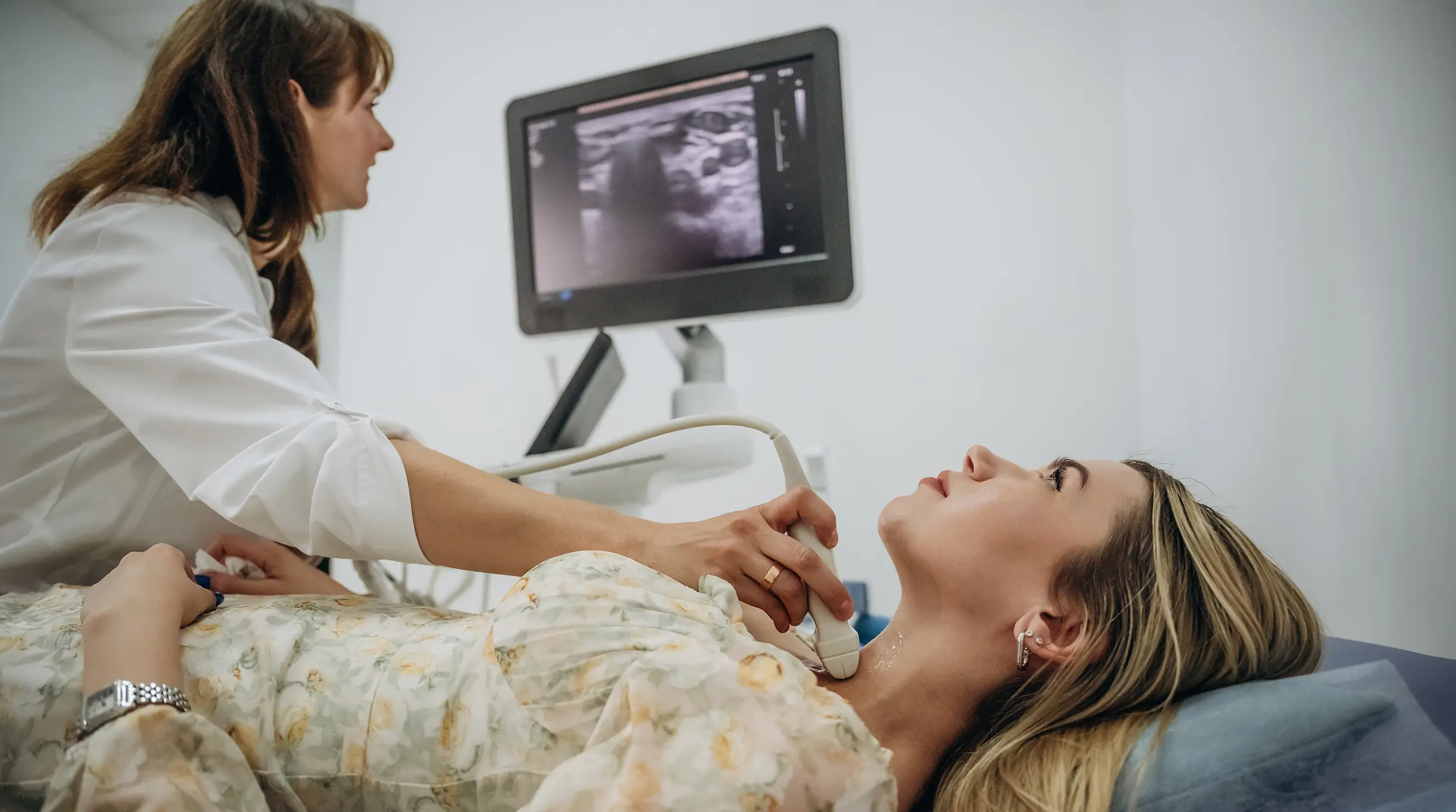 A woman lies on an examination table as an ultrasound is performed on her throat, with medical equipment in view.